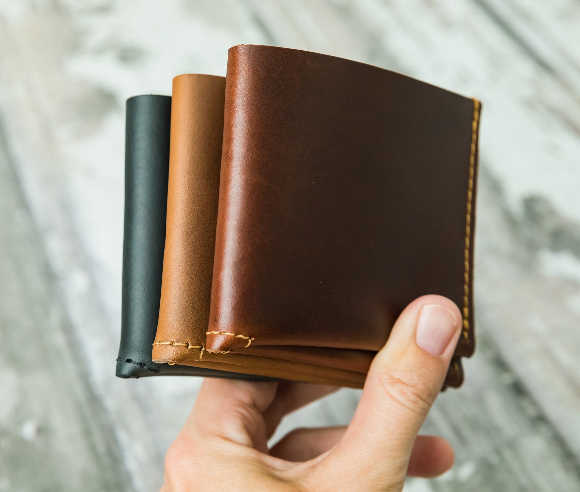 Three leather wallets in black, brown, and another brown shade held by a hand against a blurred natural background. personalized gift for dad